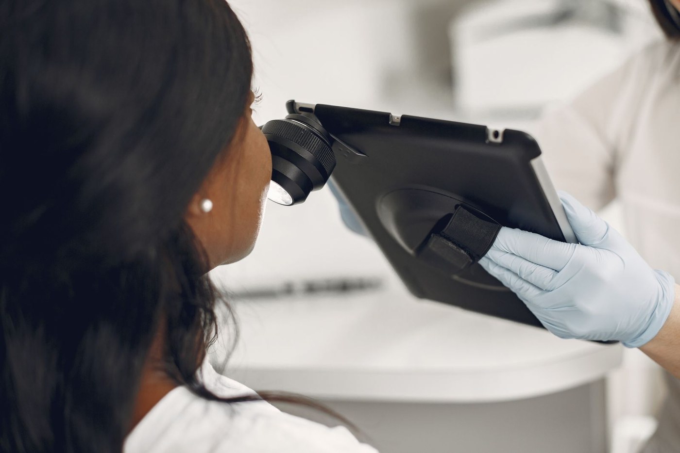 Examen de la peau au dermatoscope : le praticien, ganté, applique la loupe éclairante reliée à une tablette contre la joue d'une patiente